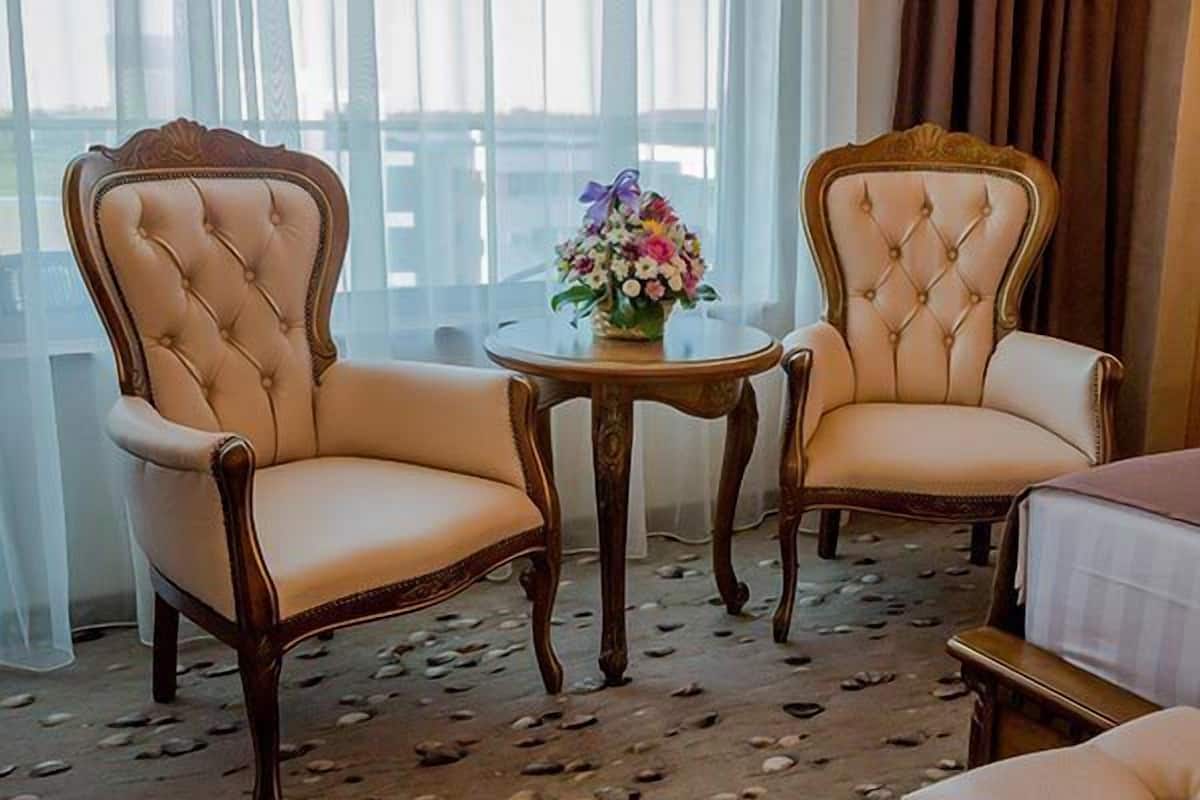 A hotel room with two upholstered chairs, a small round table with a floral centerpiece, and a bed with a white bedspread.