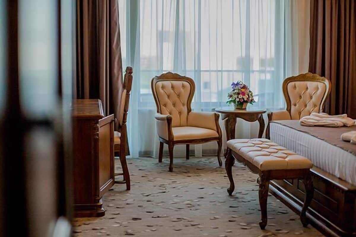 A hotel room with a bed, two upholstered chairs, a small round table with a vase of flowers, and a bench.
