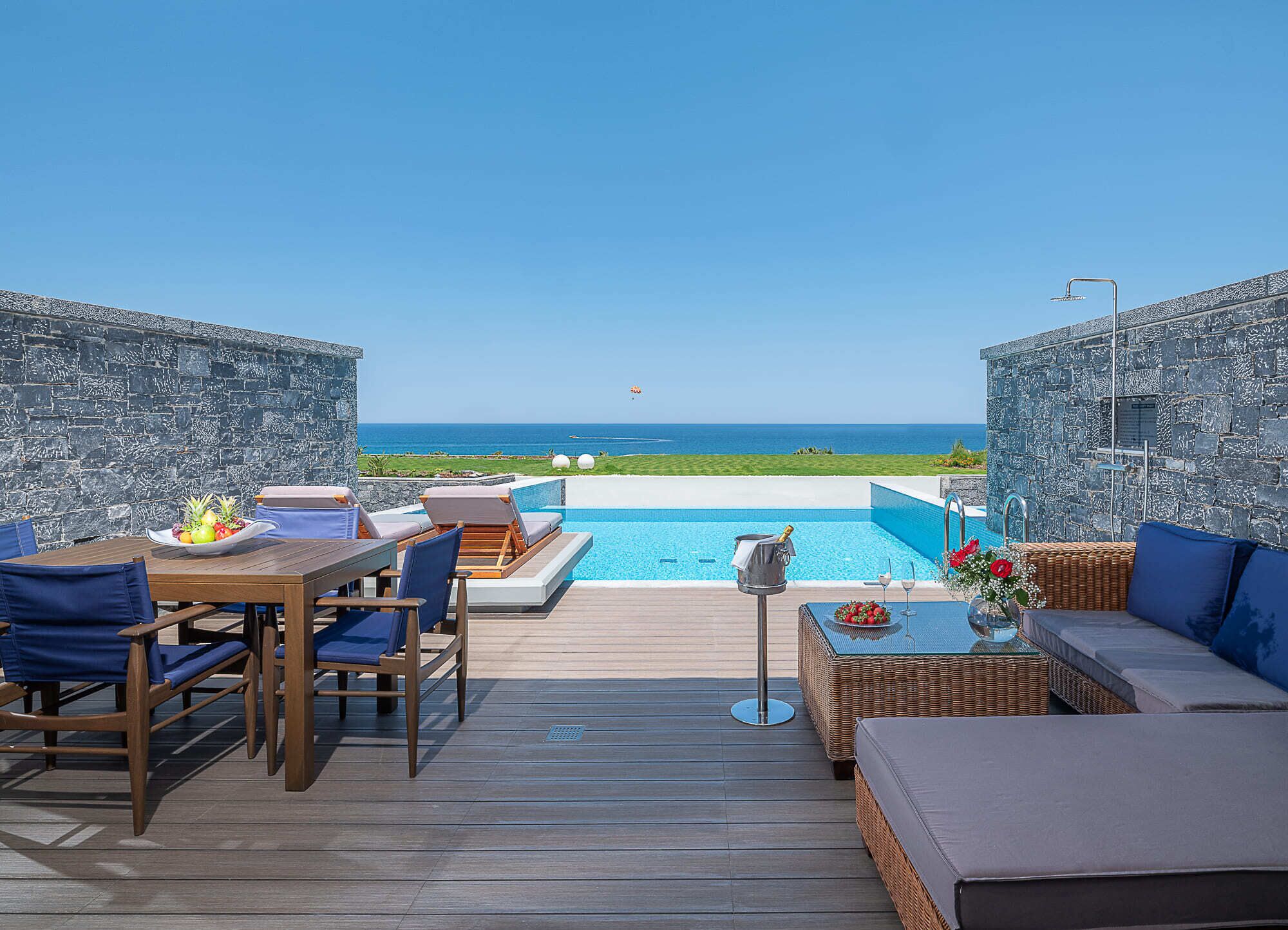 A poolside area with a view of the sea, wooden furniture, and a stone wall.