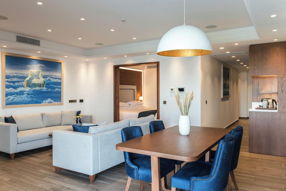 A modern hotel room with a dining area, a sofa, a wooden dining table, and a large painting on the wall.