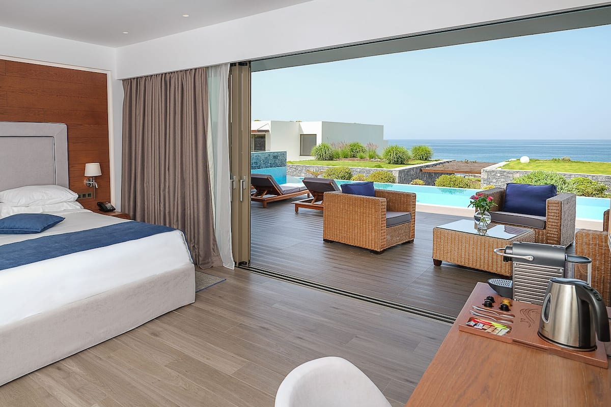 A hotel room with a bed, a desk with a kettle, and a view of the ocean through a sliding glass door.