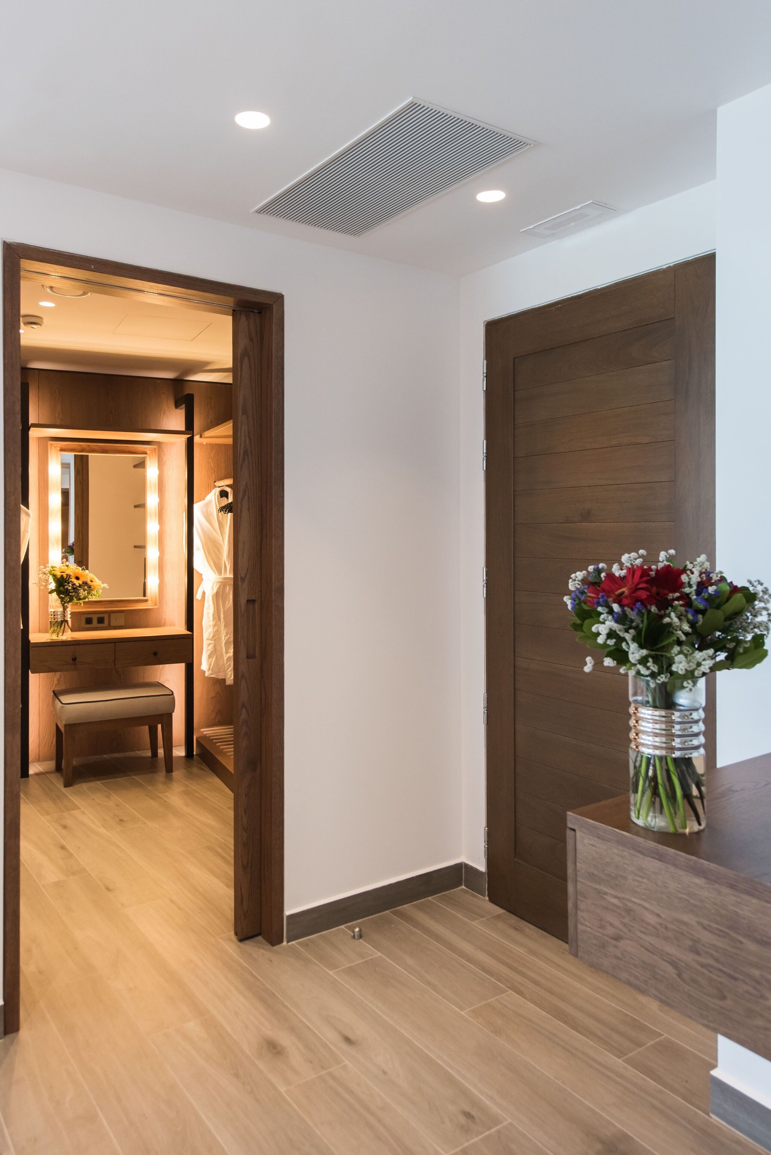 Habitación de hotel con piso de madera, puerta de madera, tocador con espejo e iluminación, un banco y un florero con flores.