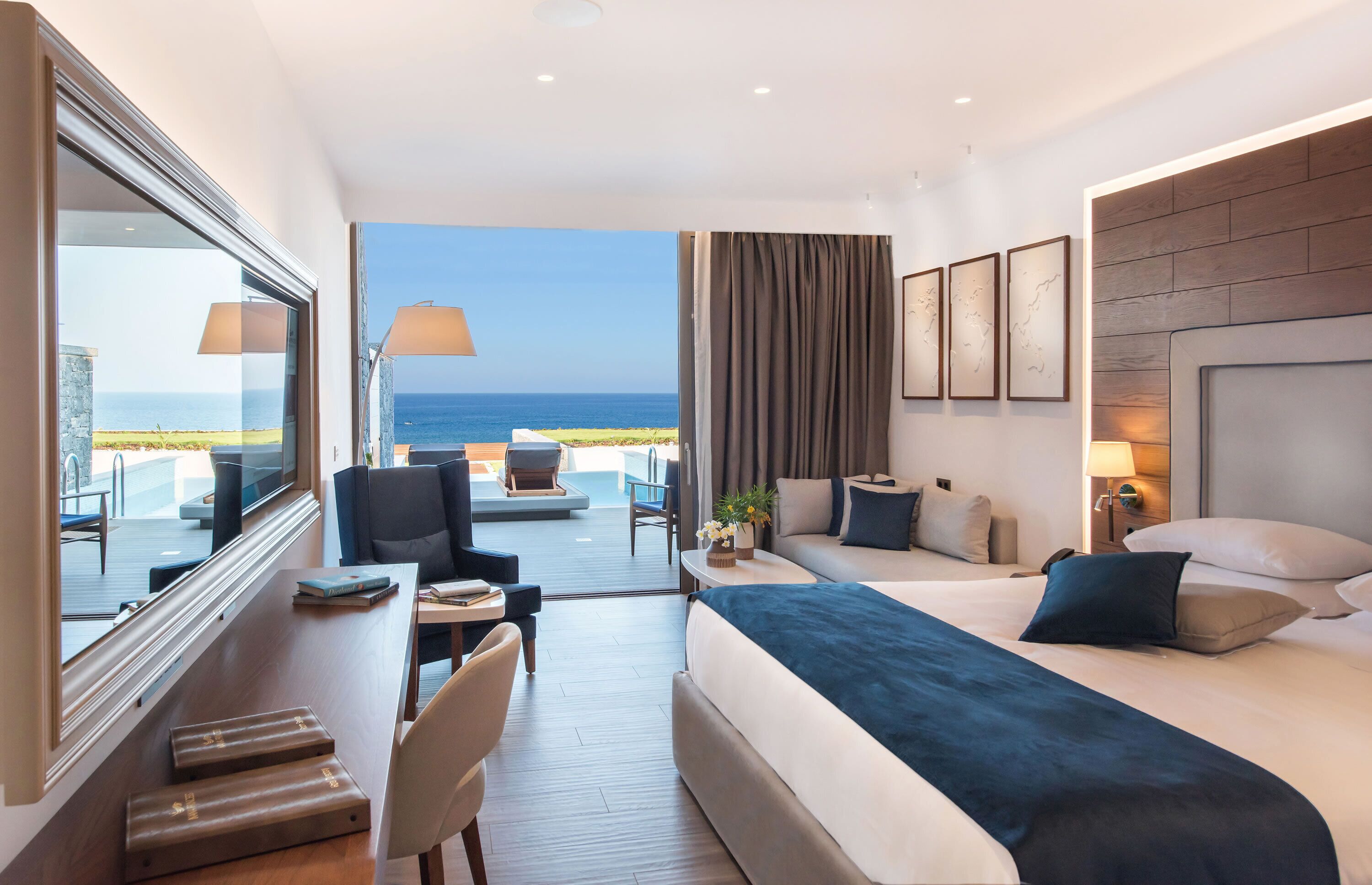 A hotel room with a bed, a desk, a chair, a sofa, a TV, and a view of the ocean.