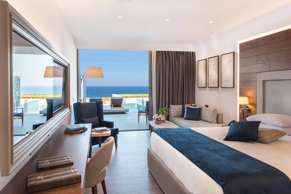 A hotel room with a bed, a desk, a chair, a sofa, a TV, and a view of the ocean.