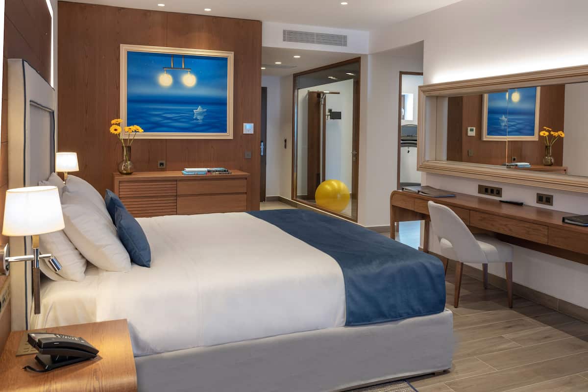 A modern hotel room with a large bed, a desk, a chair, a desk lamp, a phone, a mirror, and a painting of a night scene.