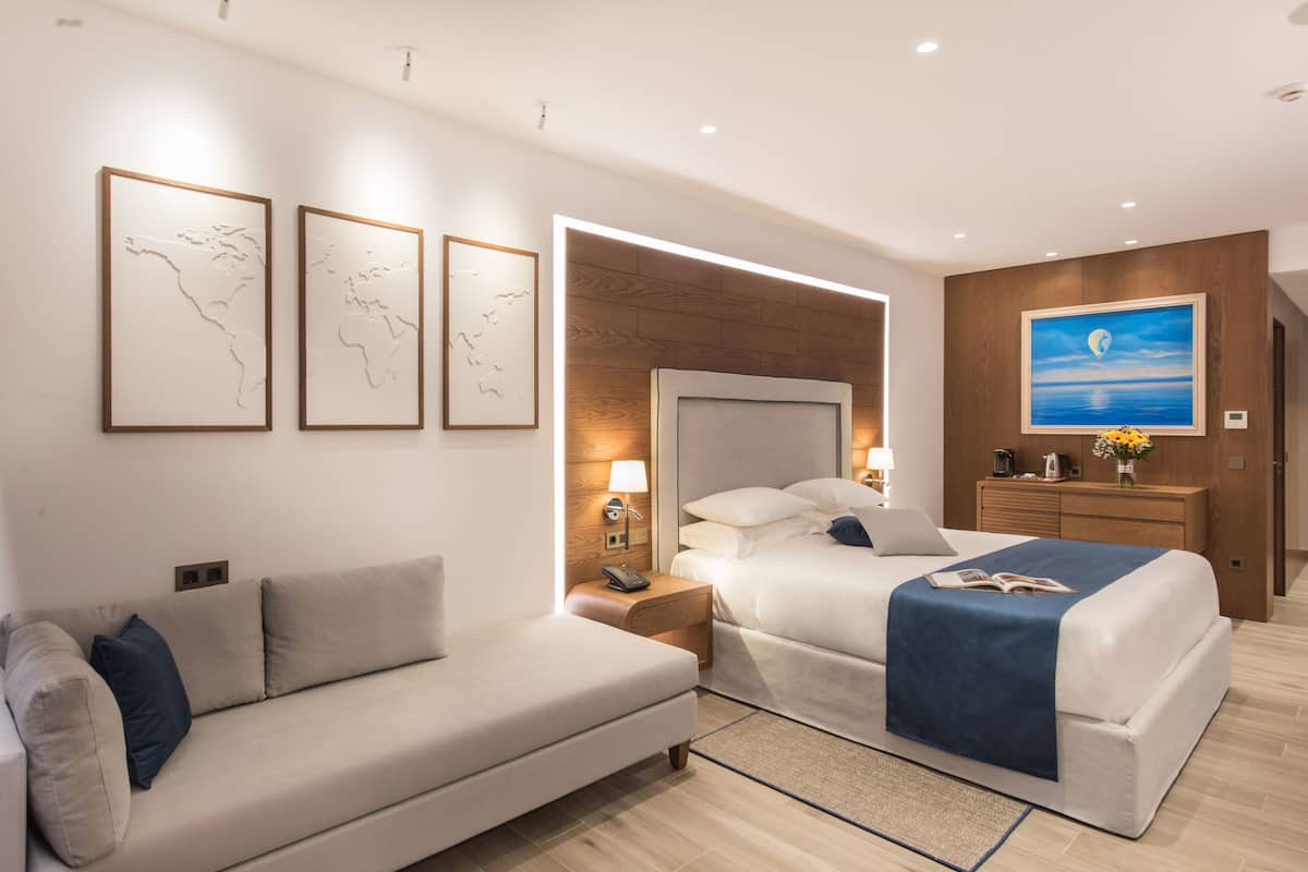 A modern hotel room with a large bed, a sofa, and a desk. There are framed pictures on the wall, a painting, and a wooden headboard.