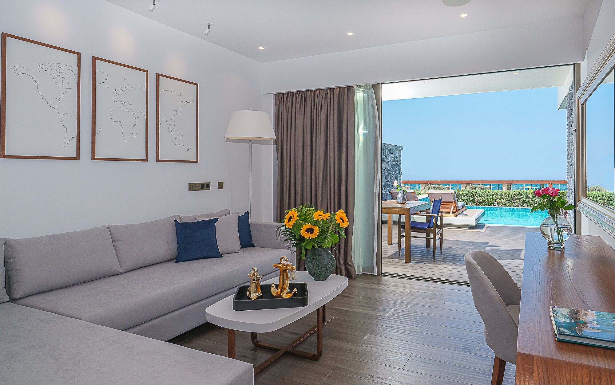 A modern hotel room with a grey sofa, a small table with a vase of sunflowers, a view of a pool and outdoor seating area.