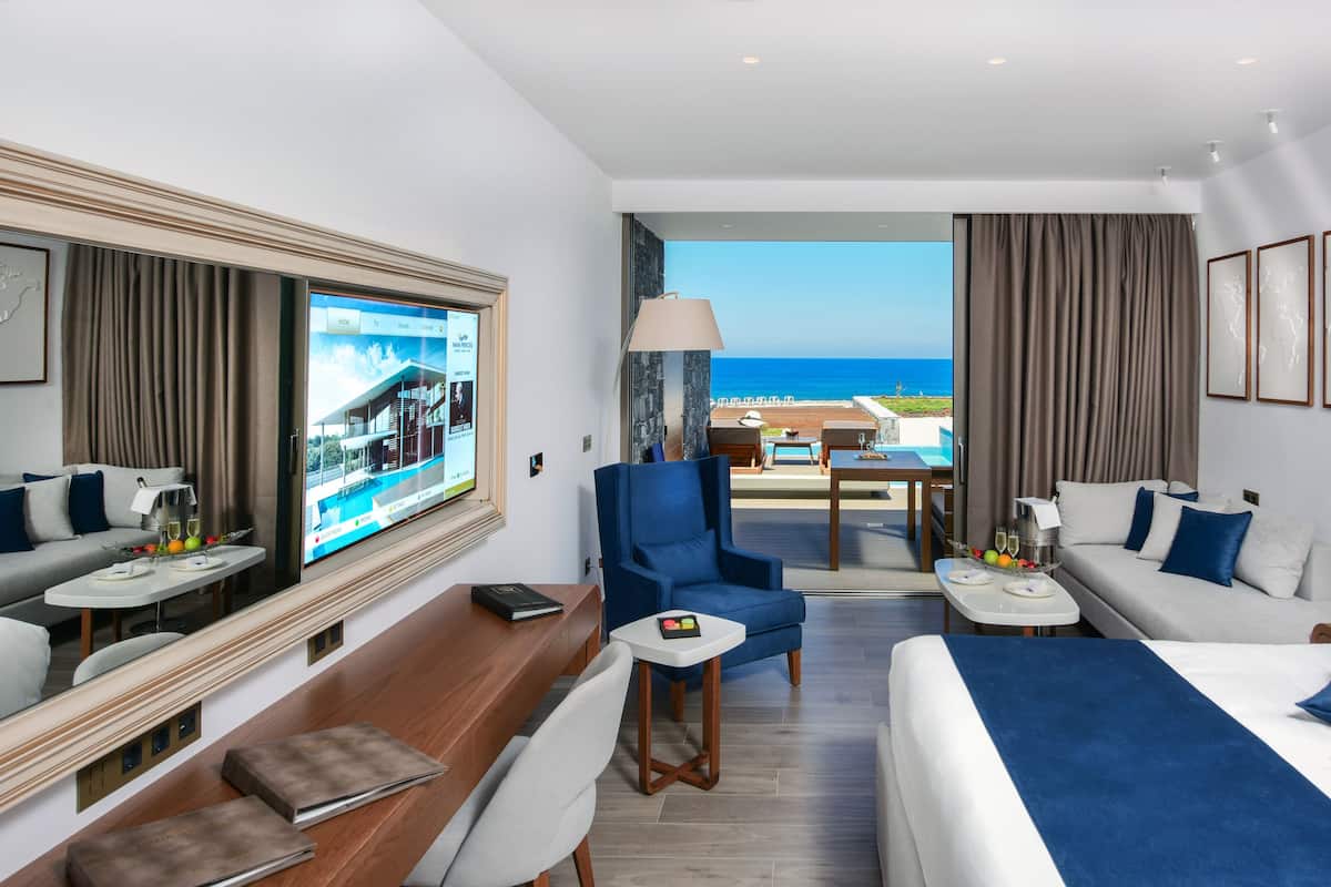 A hotel room with a large bed, a desk, a chair, a sofa, and a balcony with a view of the ocean.