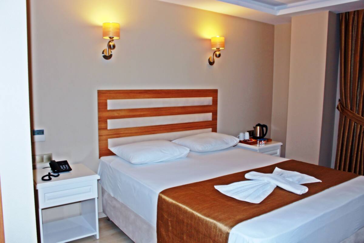 Habitación de hotel con cabecera de madera, ropa de cama blanca, dos lámparas de noche, una mesita de noche con cafetera y un teléfono.