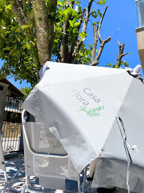 A covered outdoor seating area with a table and chairs, surrounded by trees and a building. - Residencial Casa Flora (Bombinhas)