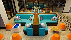 Een moderne lobby met blauwgroene banken, oranje poufs en een centrale houten tafel.