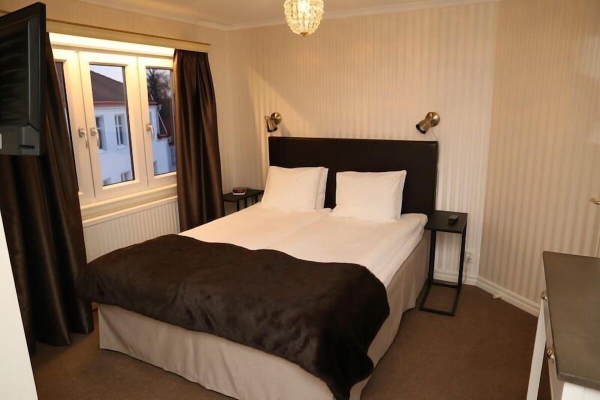 A hotel room with a bed, a nightstand, a lamp, and a window with curtains.