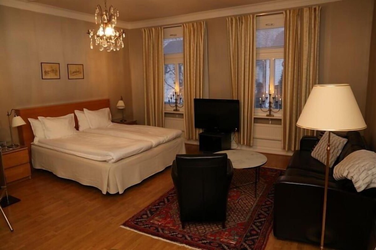 A hotel room with a large bed, a TV, a sofa, a coffee table, and a chandelier.