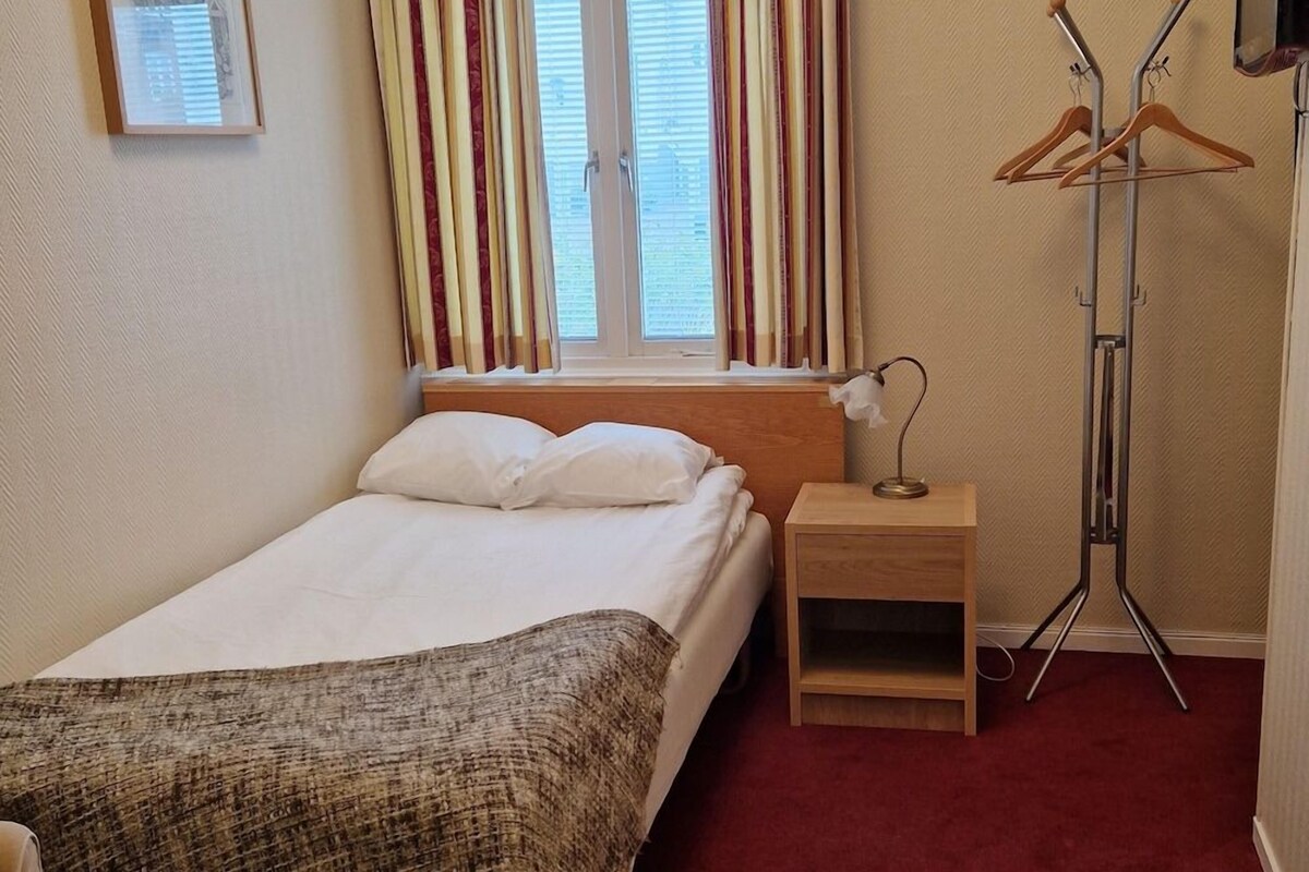 A hotel room with a bed, a bedside table, a lamp, a coat rack, and a window with curtains.