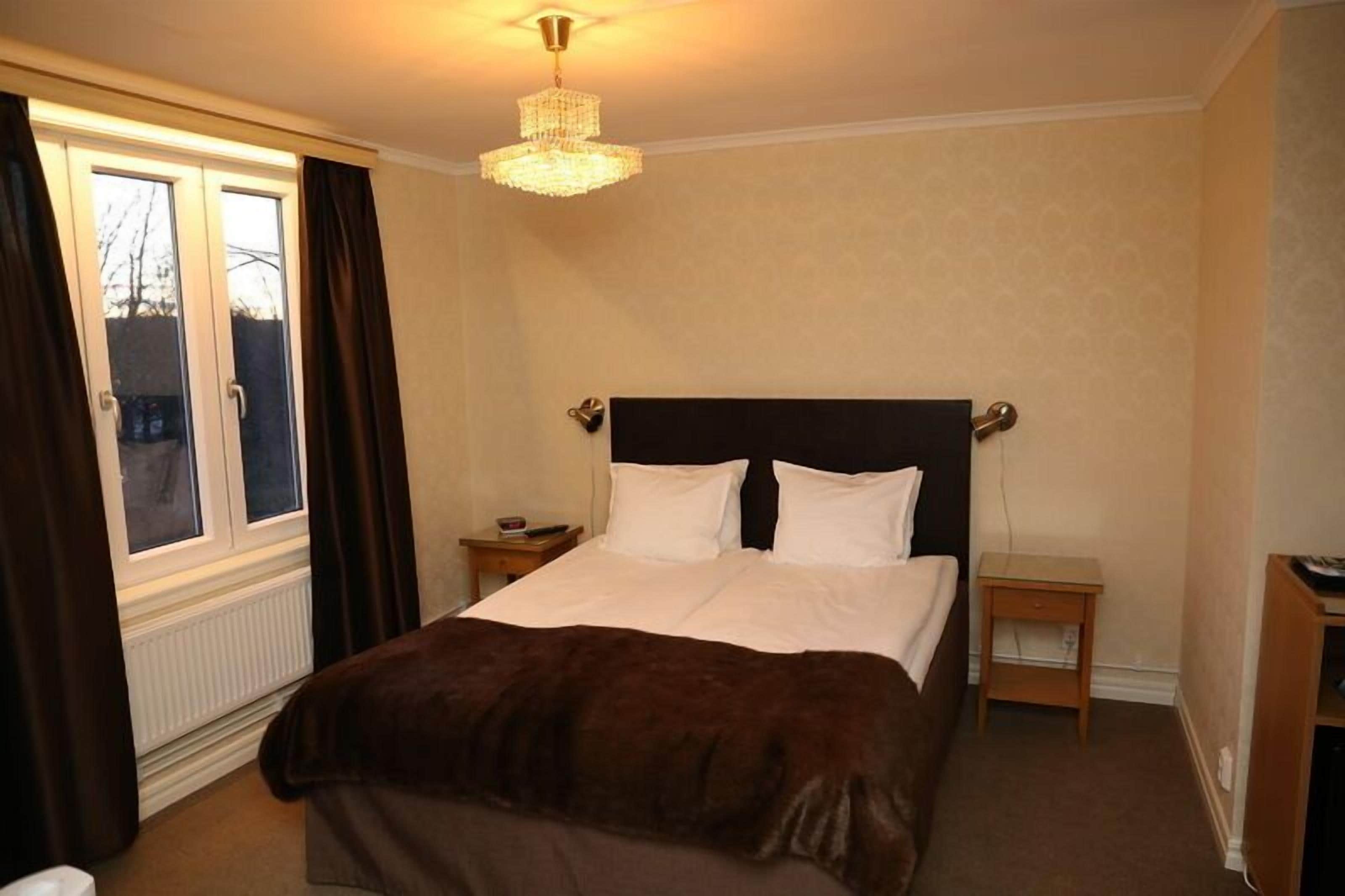 A bedroom with a large bed, a nightstand, a dresser, a window with curtains, and a chandelier.