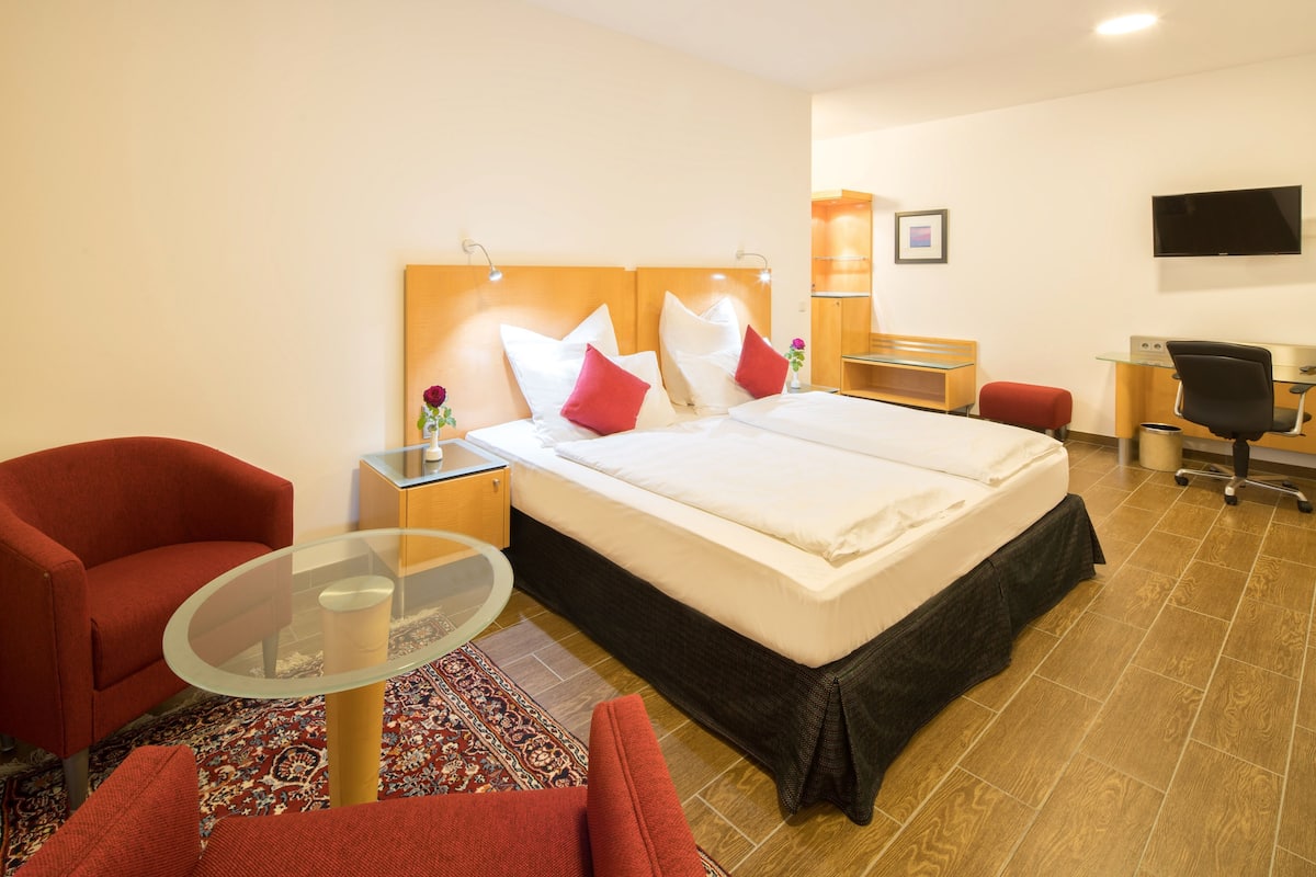 Habitación de hotel con una cama grande, dos sillones rojos, una mesa de vidrio, un escritorio con una silla, un televisor y una alfombra pequeña.