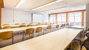 Un aula con mesas largas y sillas, una pizarra y ventanas con persianas.