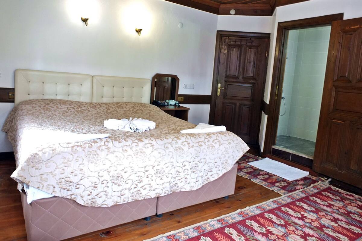 A neatly made bed with a patterned bedspread and a folded towel, a wooden nightstand with a lamp, a wooden door, and a red patterned rug on a wooden floor.