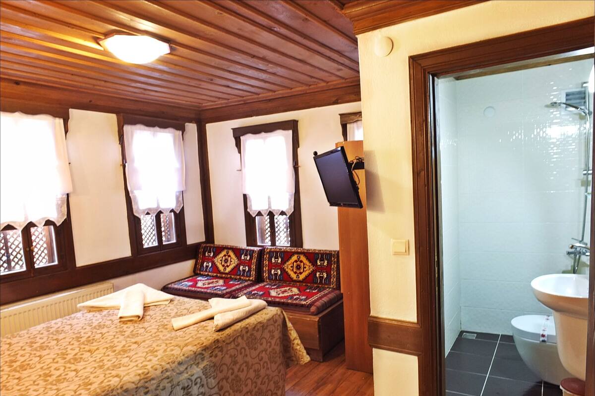 A room with a bed, a bench with patterned cushions, a television mounted on the wall, and a bathroom with a sink and toilet.