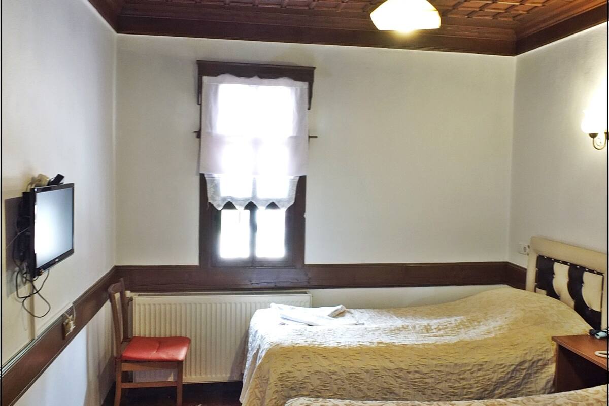 A hotel room with two beds, a wooden ceiling, a window with curtains, a radiator, a television, and a red chair.