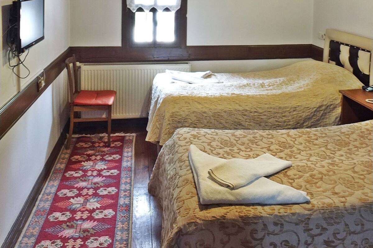 A room with two beds, a window with curtains, a radiator, a television, and a wooden ceiling.