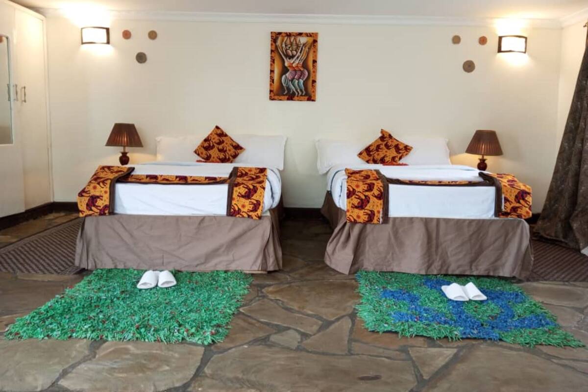 A hotel room with two beds, each with a brown bedspread and matching pillows. There are two wall-mounted lamps, a painting above the beds, and a stone floor.