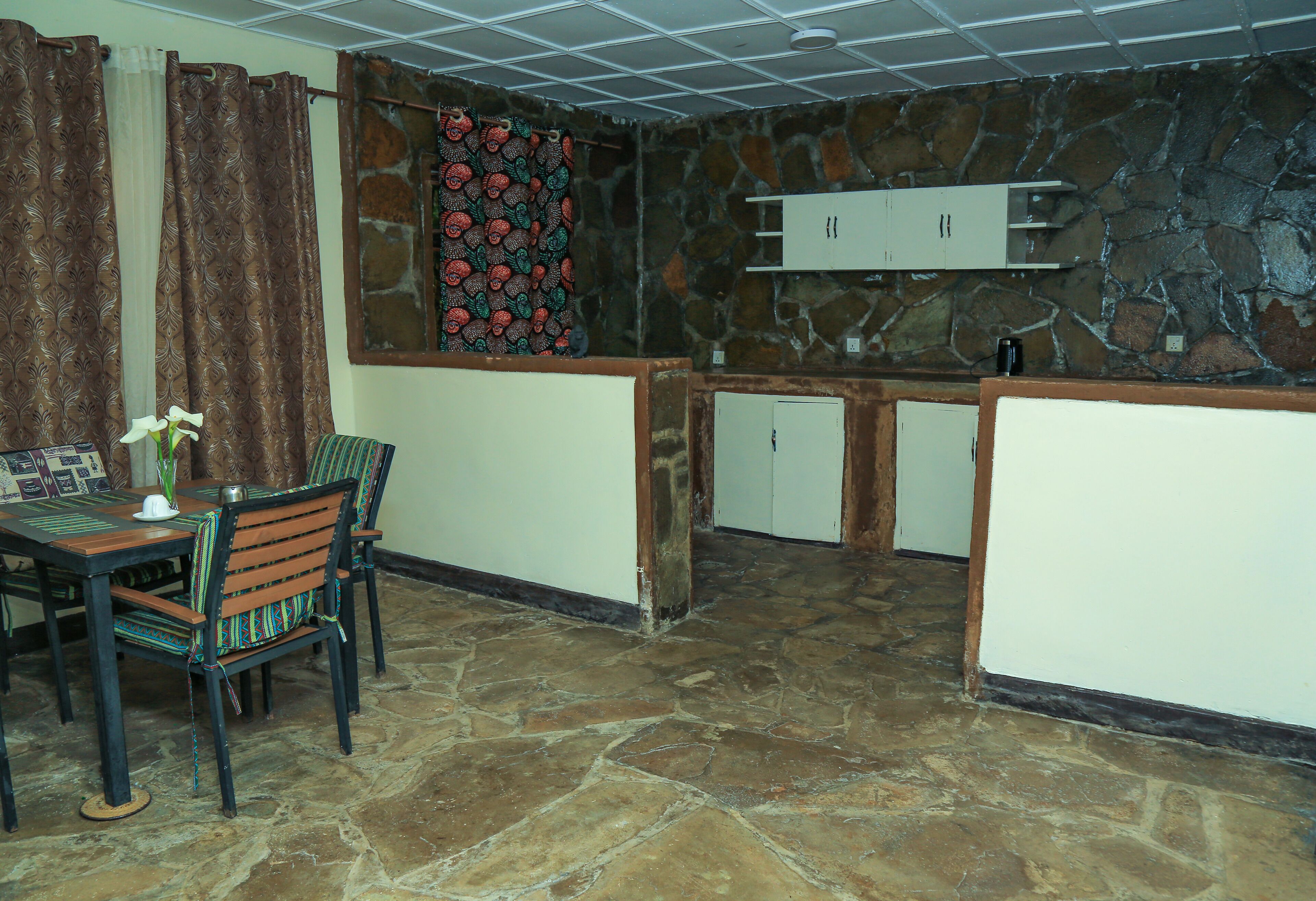 A stone-floored room with a dining table and chairs, a patterned curtain, and a stone wall with a decorative hanging.