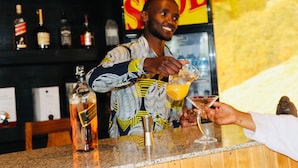 Un cantinero sirve una bebida en una copa de martini en un bar con mostrador de madera y estantes llenos de botellas de licor.