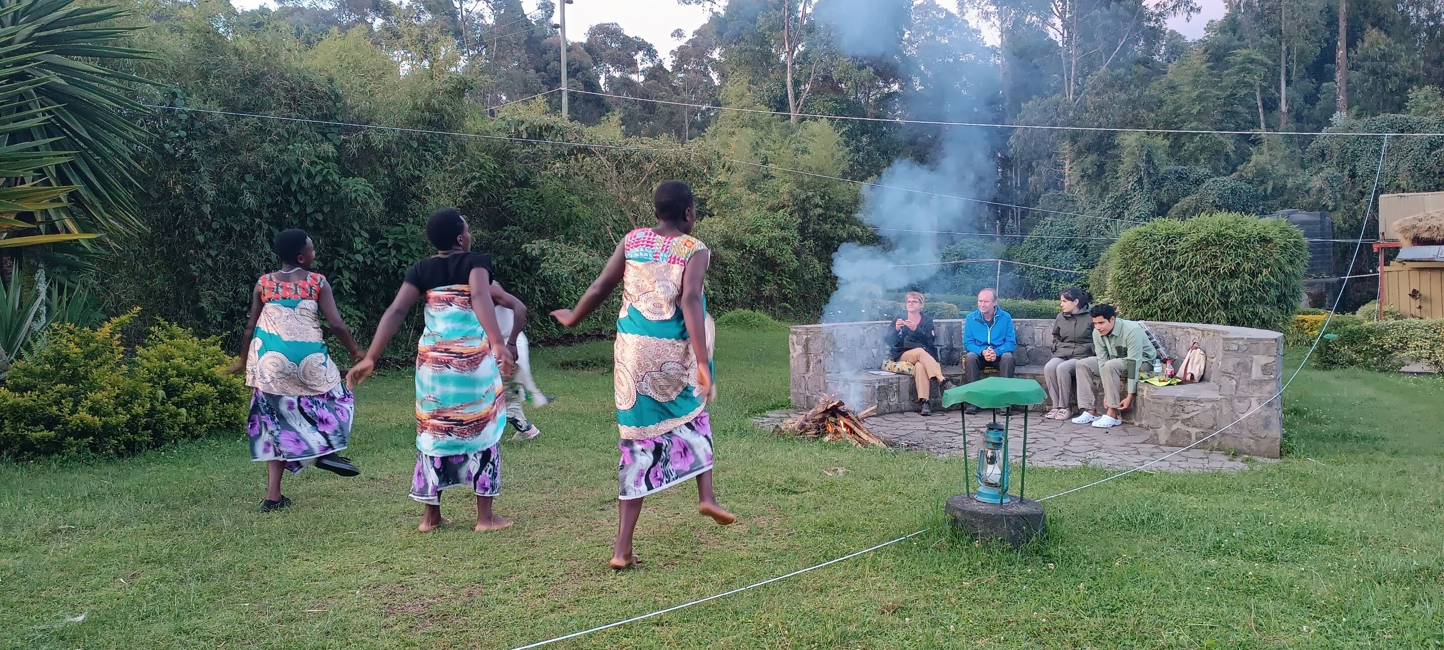 Un grupo de personas reunidas alrededor de un hogar de piedra en un espacio al aire libre cubierto de césped, con abundante vegetación al fondo.