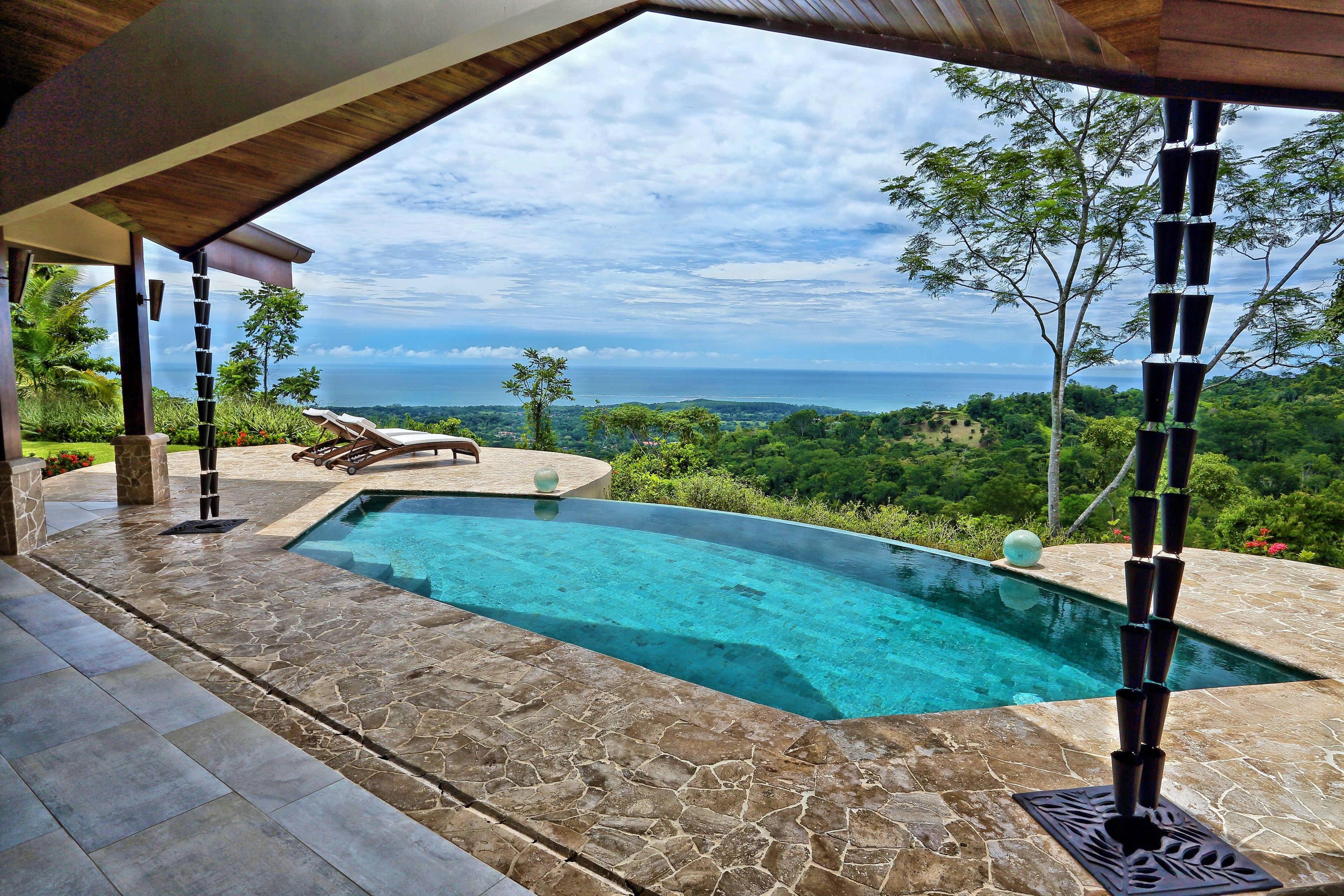A swimming pool with a clear view of the ocean, surrounded by stone tiles and a wooden pergola.