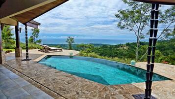 A swimming pool with a clear view of the ocean, surrounded by stone tiles and a wooden pergola.
