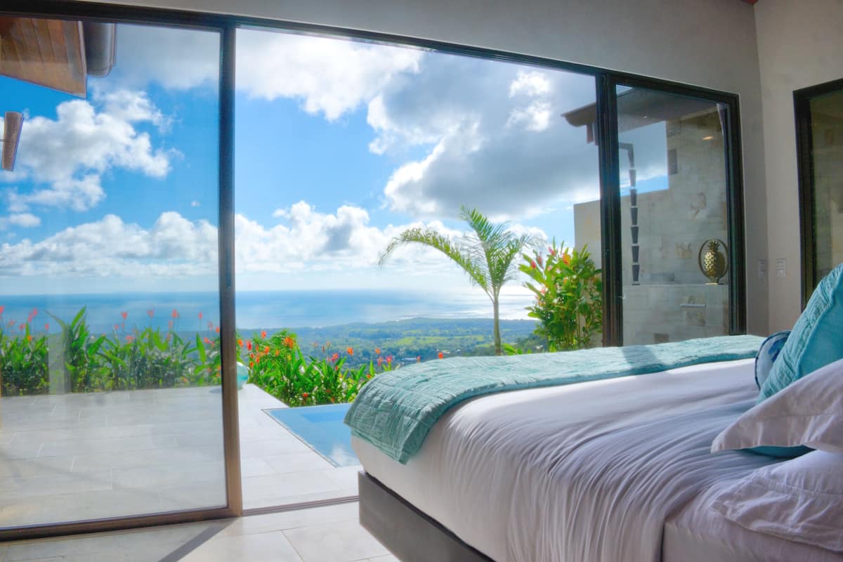 A bedroom with a large sliding glass door leading to a balcony with a view of the ocean and hills.