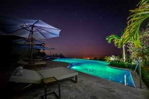 Un área de piscina nocturna con sillones de descanso y sombrillas, rodeada de suelo de piedra y vegetación.