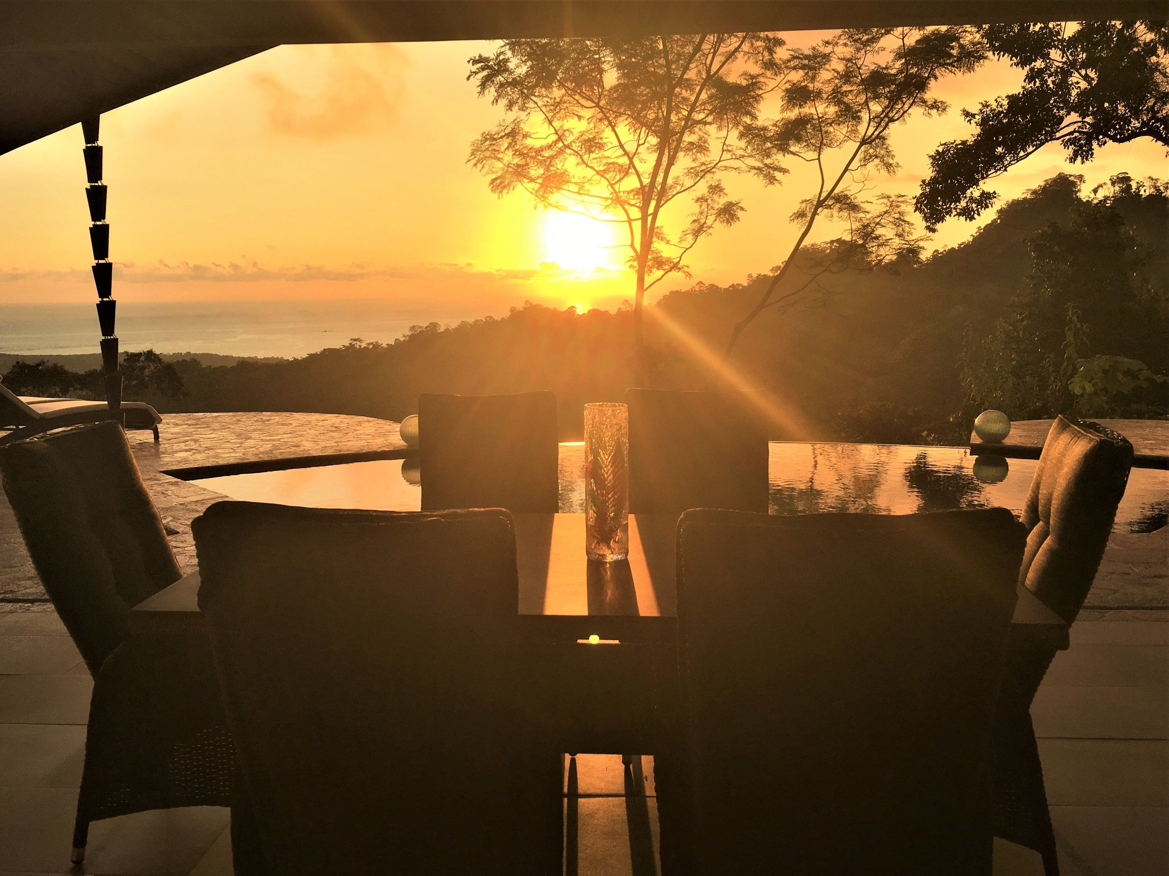 A dining area with a view of the sunset over the sea, outdoor furniture, and a water feature.