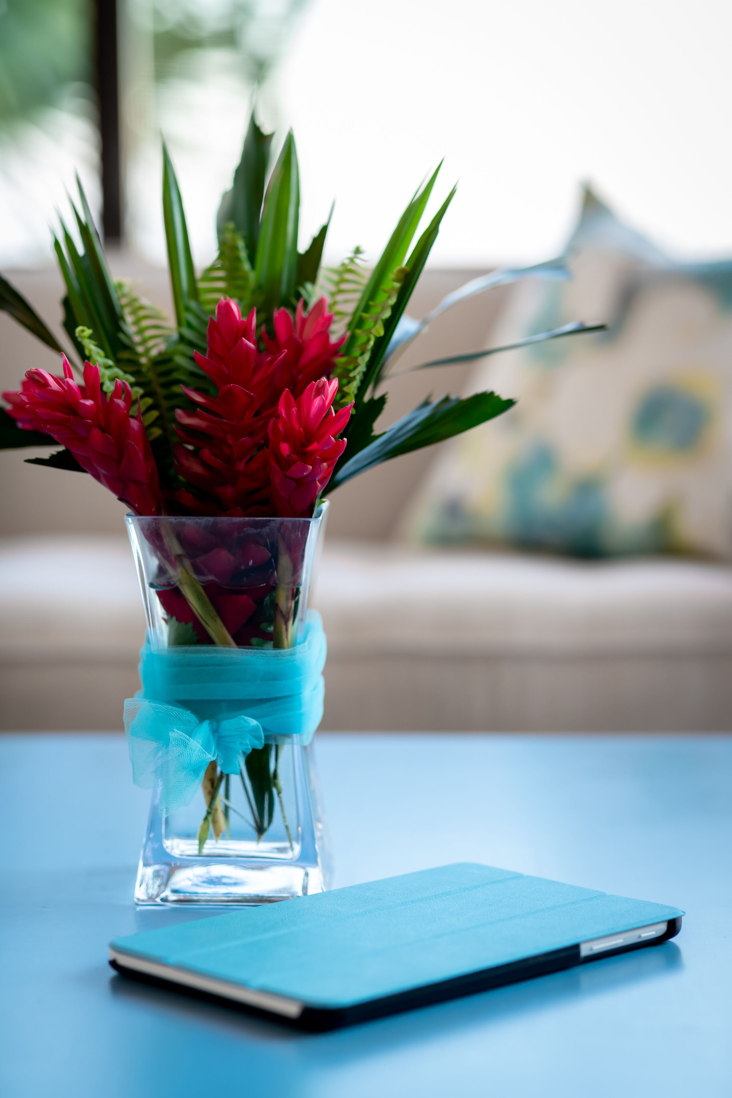 Eine klare Glasvase mit roten Blumen und grünen Blättern auf einem blauen Tisch.