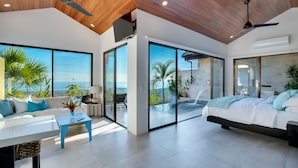 A modern bedroom with a large bed, a sofa, and a dining table. It features sliding glass doors leading to a balcony with a view of the ocean. - Golden Pineapple Villas (Bahía Ballena)