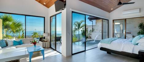 Un dormitorio moderno con una cama grande, un sofá y una mesa de comedor. Cuenta con puertas de cristal correderas que dan a un balcón con vistas al océano.