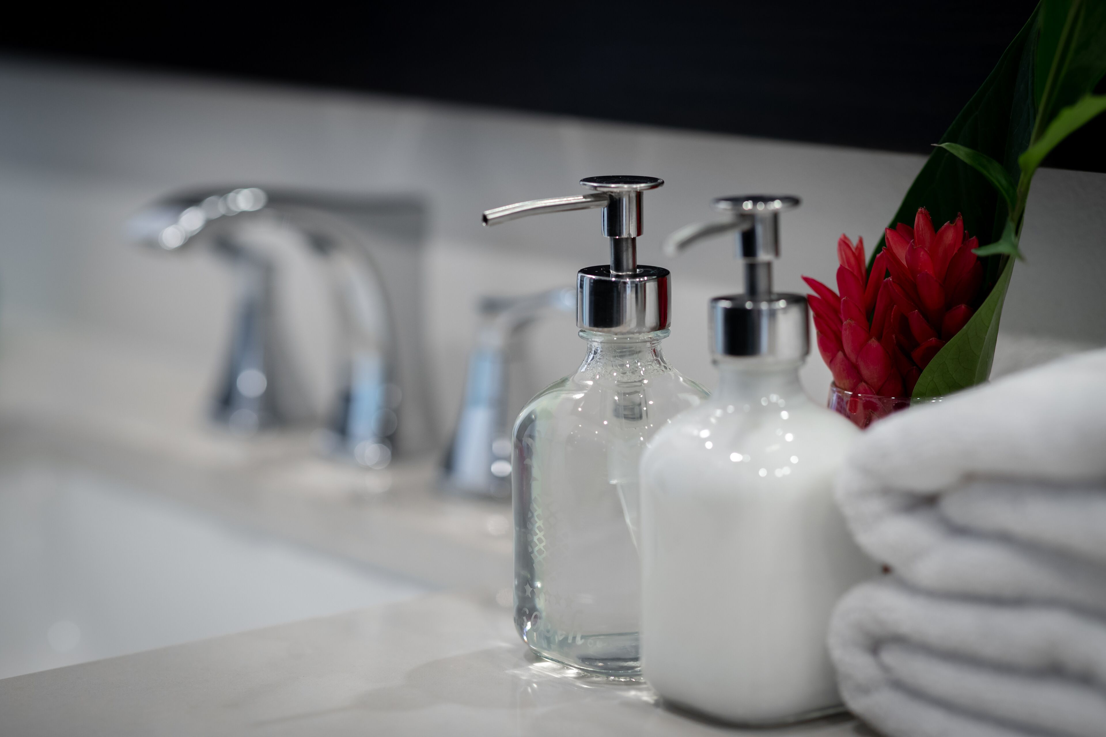 A bathroom with a modern faucet, two clear glass soap dispensers, and a red flower in a vase.