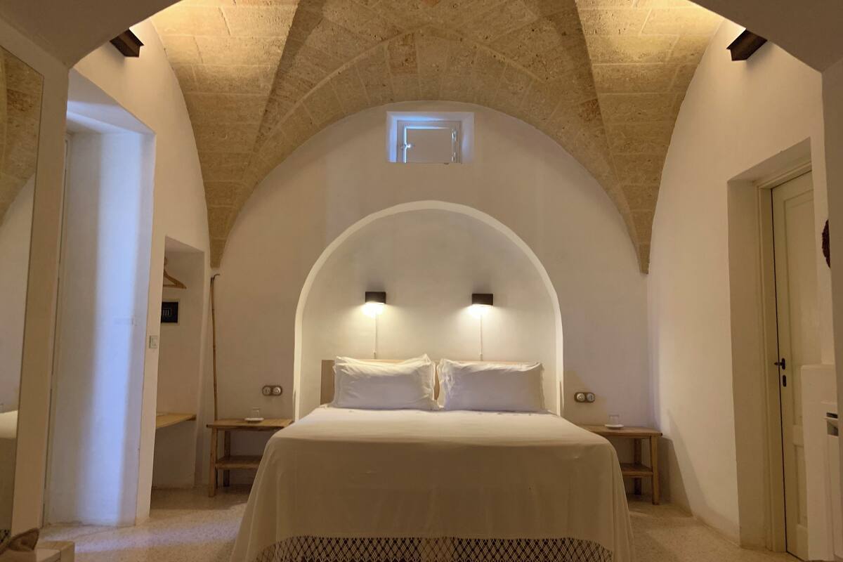 A bedroom with a vaulted ceiling, a large bed, bedside tables, and a small window.