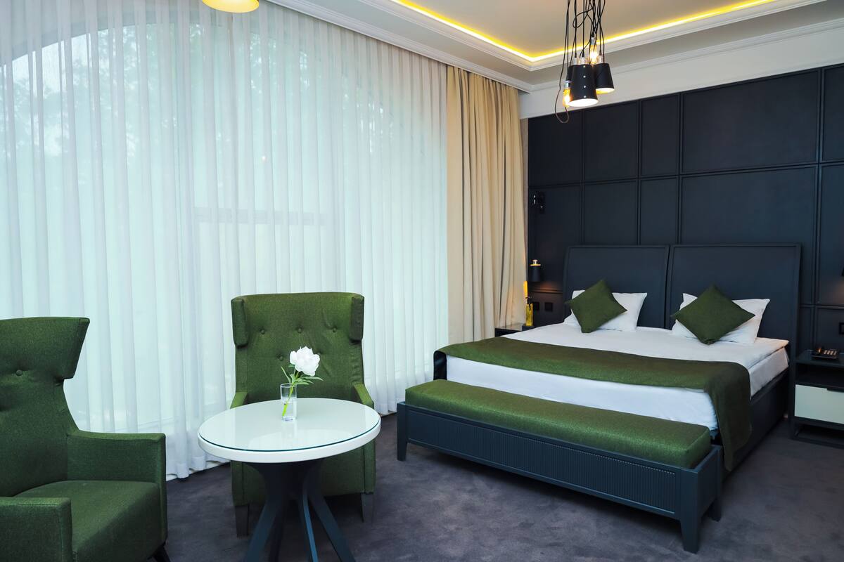 A hotel room with a large bed, two green armchairs, a small round table with a vase, and a dark headboard.