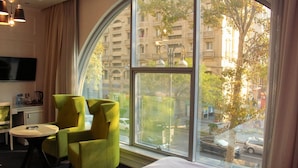 Una habitación de hotel con un gran ventanal de arco, una cama, un sillón verde, una mesita redonda y un televisor de pantalla plana montado en la pared.