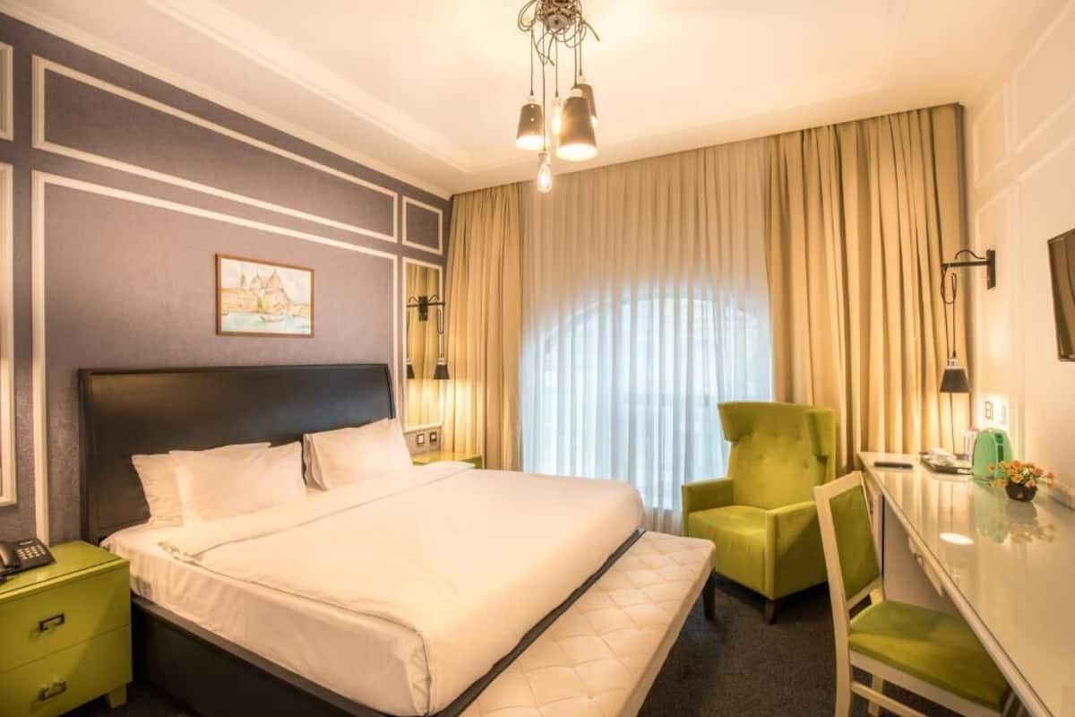 A hotel room with a large bed, a desk with a chair, a green armchair, and a painting on the wall.