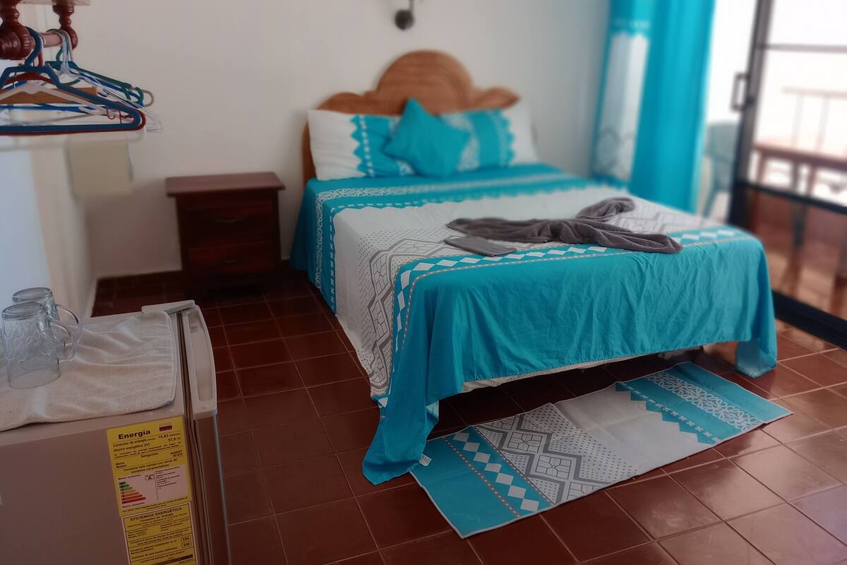 Een slaapkamer met een bed, een nachtkastje, een houten dressoir, een wandlamp en een raam met gordijnen.