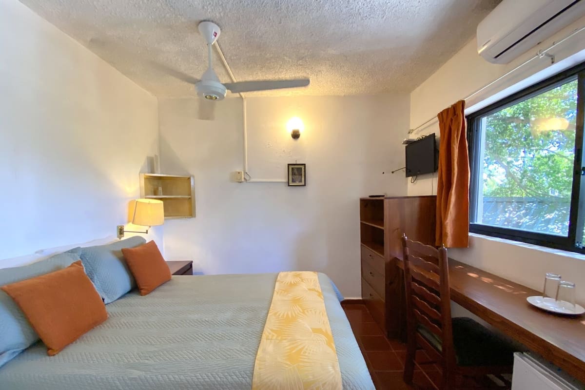 Een slaapkamer met een bed, een bureau, een stoel, een airconditioner, een raam met gordijnen en een tv aan de muur.