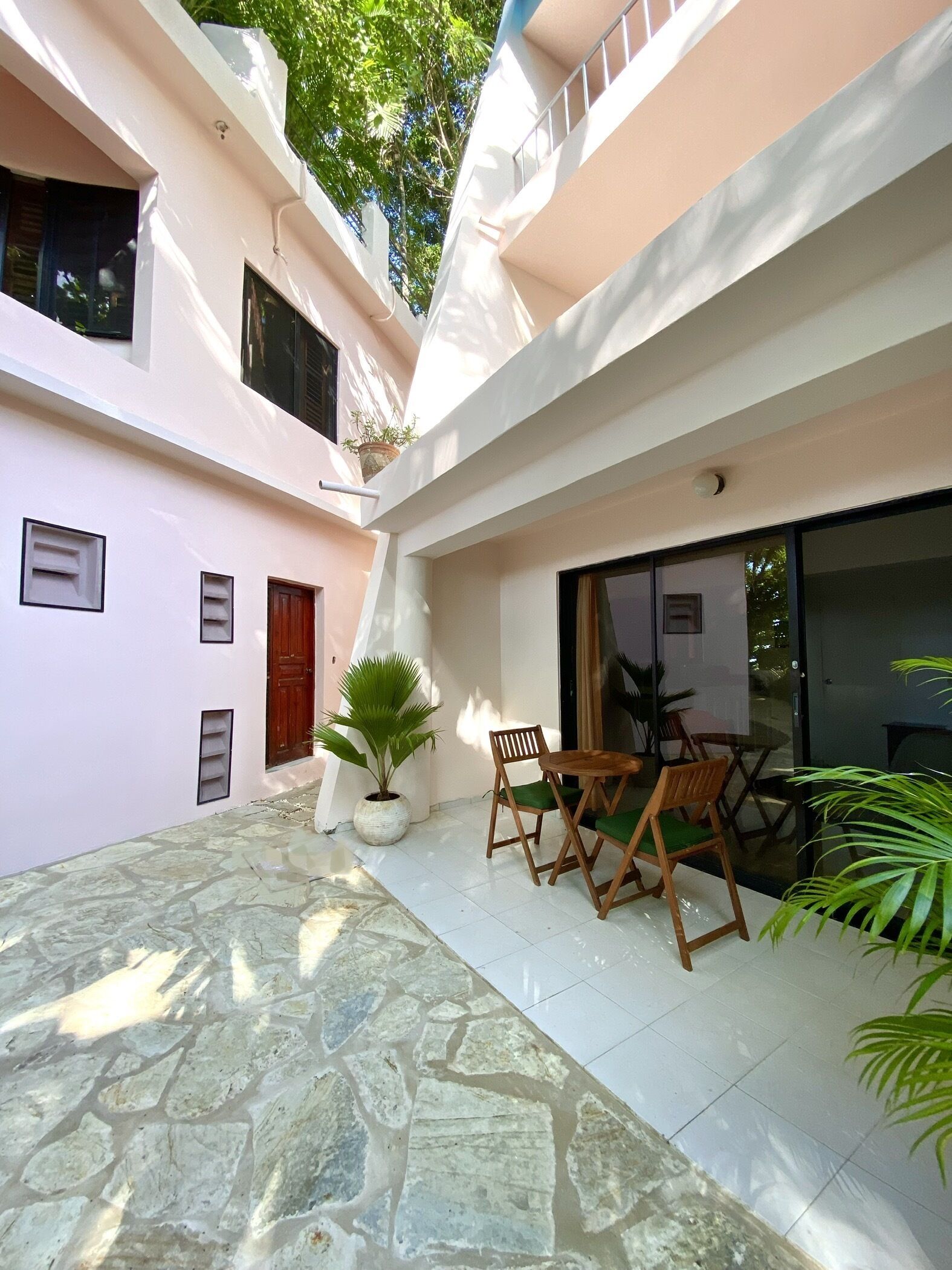 A courtyard with a stone floor, a wooden table with chairs, potted plants, and a building with a pink facade.