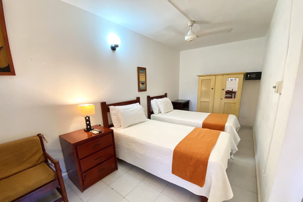 Een hotelkamer met twee bedden, een houten nachtkastje, een stoel, een kledingkast en een plafondventilator.