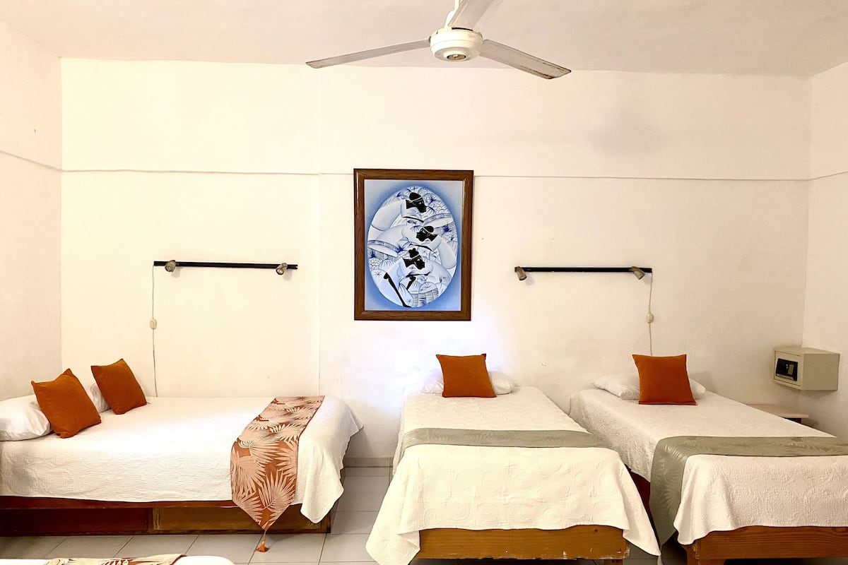 Een hotelkamer met drie eenpersoonsbedden, elk met een witte spreidek en oranje kussens. Er is een plafondventilator, een lijstwerk met een kunstwerk boven de bedden, en een klein nachtkastje met een lamp.
