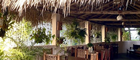 An outdoor dining area with thatched roof, wooden tables, and chairs, surrounded by potted plants and palm trees.