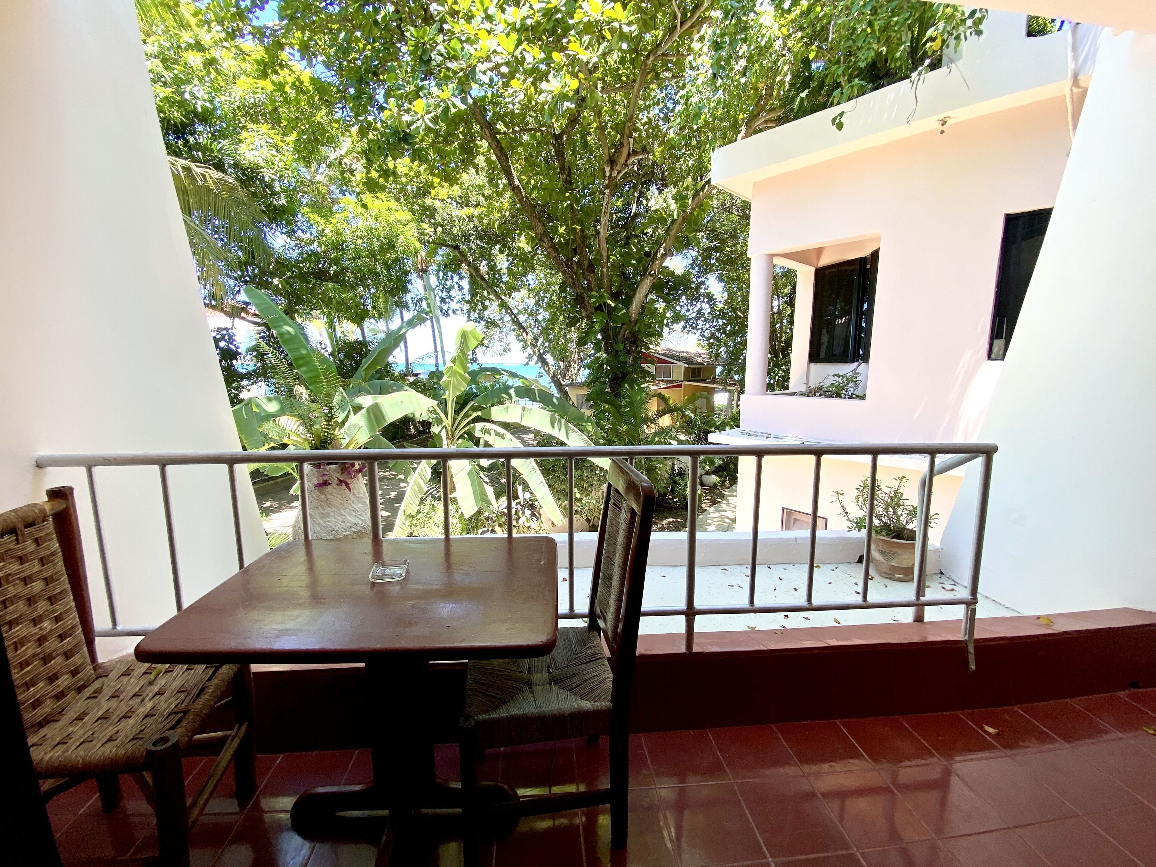 A balcony with a table and chairs, surrounded by a railing, overlooking a garden and a building.