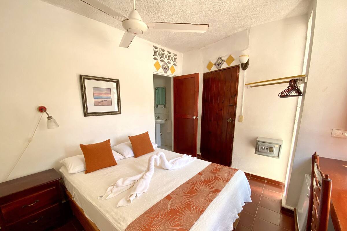 Hotelkamer met een bed, nachtkastje, dressoir, plafondventilator, wandlamp en deur naar een badkamer.